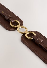 Bracelet en cuir marron avec quincaillerie métallique dorée, doté d'une connexion en forme de boucle ovale, bordée de coutures contrastantes et de surfaces lisses.