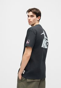 Νεαρός με κοντά σκούρα μαλλιά, ντυμένος με μαύρο μπλουζάκι Nike με μεγάλο λογότυπο στην πλάτη και παντελόνι cargo, ποζάρει πλαγίως με τα χέρια στις τσέπες.