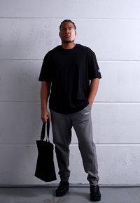 Maglietta nera, pantaloni della tuta grigi con logo e borsa tote nera; in piedi contro uno sfondo grigio uniforme, indossando scarpe nere.
