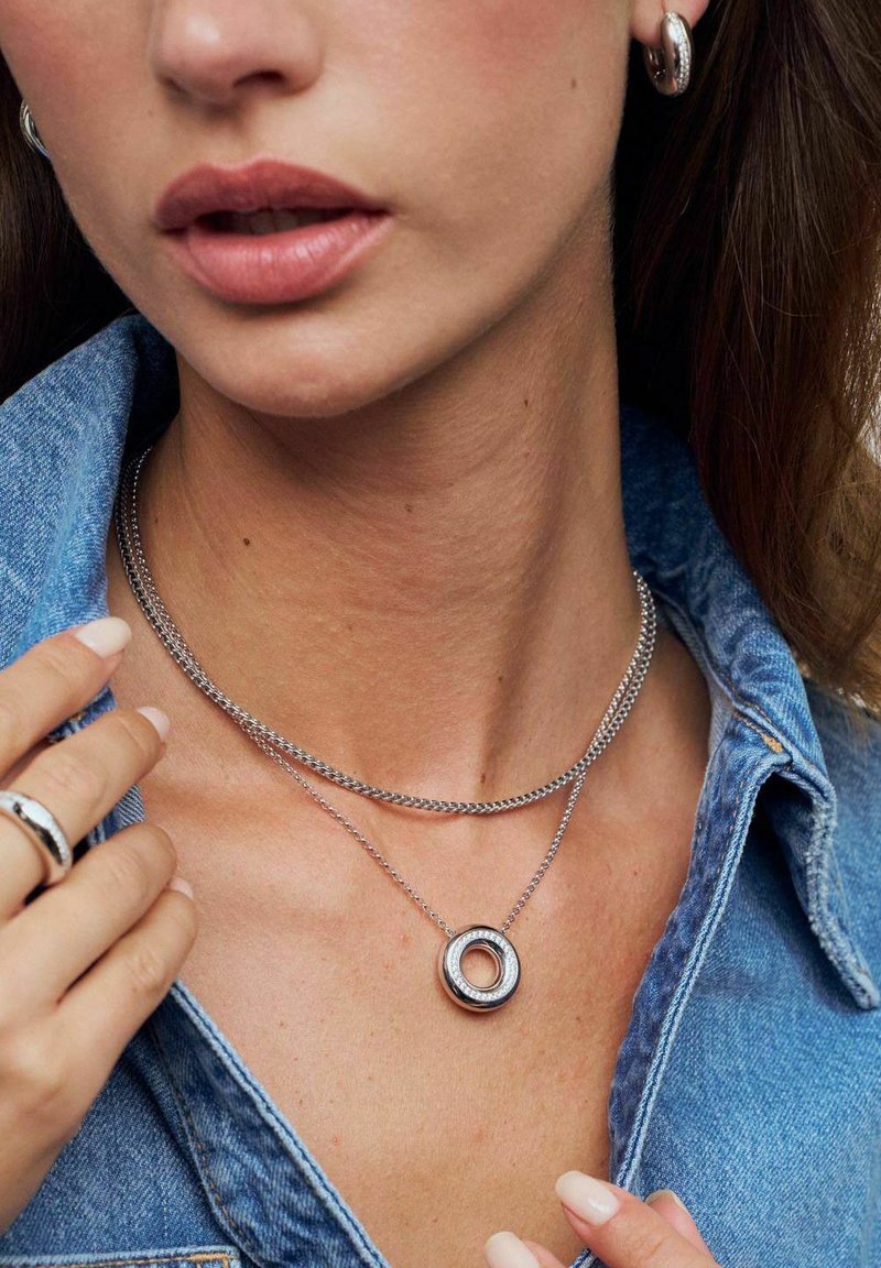 Silversmycken i kedjor som är lager på lager runt halsen, med en cirkulär medaljong med en glänsande yta och en liten klar detalj.