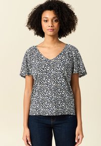 Top floreale a V in navy con motivi bianchi e azzurri. Maniche corte, tessuto morbido, vestibilità comoda, abbinato a jeans in denim scuro.