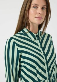 Zeleno-krémová pruhovaná košile s límečkem, knoflíkovou légou a dlouhými rukávy. Hladká látka s vertikálními a horizontálními pruhy.