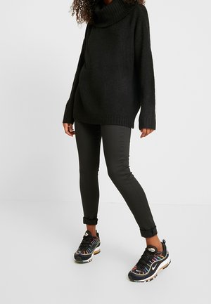 Femme portant un pull à col roulé noir, un pantalon noir ajusté et des baskets noires avec des détails colorés, debout devant un fond uni.