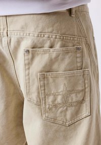 Pantalon en toile beige avec deux poches arrière, dont une avec un motif d'étoile cousue et des détails de rivets, porté avec une chemise blanche rentrée.