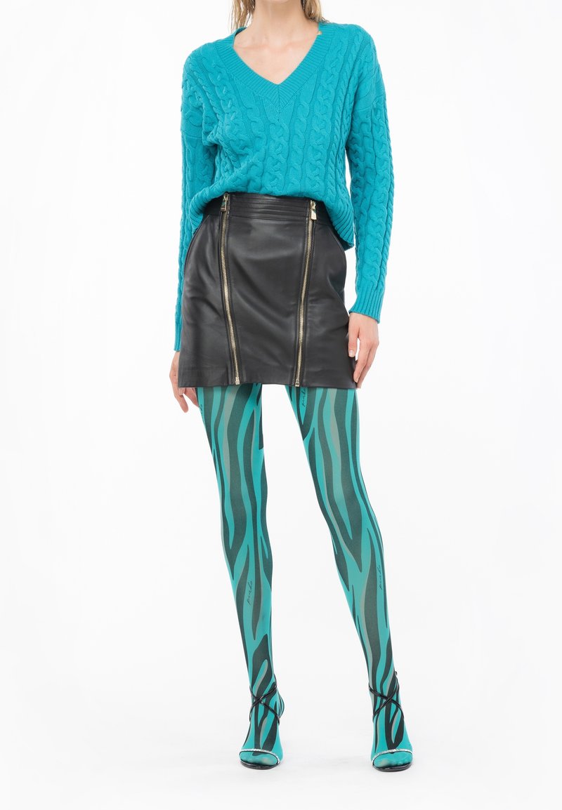 Femme portant un pull torsadé turquoise, une mini-jupe en cuir noire avec des zips, et des collants turquoise à motifs avec des chaussures assorties.