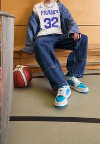 Trampki z niebieskimi i szarymi akcentami, niebieska kurtka dżinsowa, biała koszulka z napisem "FRANCE 32" oraz luźne niebieskie jeansy, siedzące obok piłki do koszykówki.
