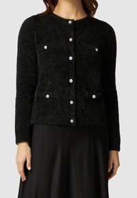 Svart fluffig cardigan med långa ärmar, rund hals och sex knäppningar med rhinestones. Två bröstfickor synliga.