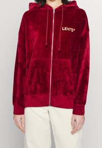 Felpa rossa in velluto con zip, con cordini, tasche frontali e logo ricamato. Tessuto spesso con una texture morbida. Polsini e orlo a coste.