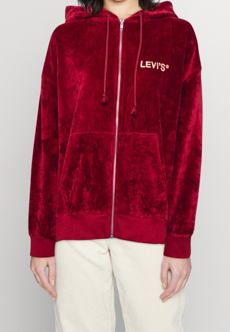 Felpa rossa in velluto con zip, con cordini, tasche frontali e logo ricamato. Tessuto spesso con una texture morbida. Polsini e orlo a coste.