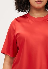 A person with curly hair wears a bright red, loose-fitting satin t-shirt, showcasing its smooth texture and casual style.