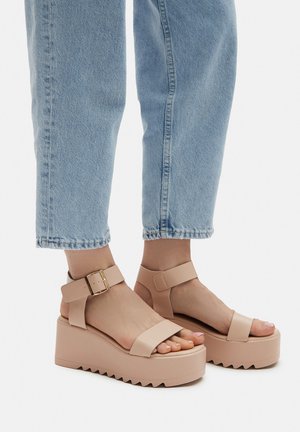 Beżowe sandały na platformie z grubą, teksturowaną podeszwą. Posiadają paski wokół kostki z złotą klamrą oraz szerokie paski na palcach. Noszone z jasnymi jeansami.