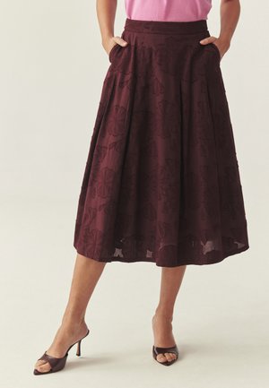 Kobieta w ciemnoczerwonej, midi spódnicy w kwiatowe wzory, różowej bluzce oraz ciemnych sandałach na obcasie z odkrytymi palcami, stoi z rękami w kieszeniach spódnicy.