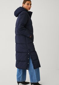Marineblauwe pufferjas met een capuchon, voorzien van een gestikte uitstraling, zijzakken en drukknoopsluitingen. Gekleed over wijde spijkerbroek en enkellaarsjes.
