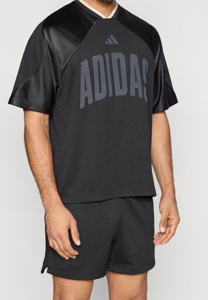 Mężczyzna noszący czarną sportową siatkową koszulkę Adidas z dopasowanymi szortami, stojący z rękami swobodnie opuszczonymi wzdłuż ciała.