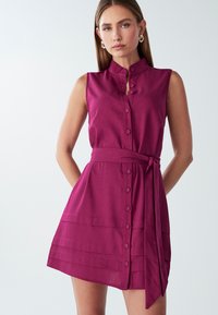 Trumpetas be rankovių, gilių magentų spalvos, su sagų apykakle, diržu nugaros linijoje ir horizontaliais plišiais sijono dalyje. Sklandi audinio tekstūra.