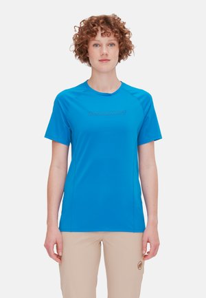 Jong persoon met krullend haar, gekleed in een felblauw Mammut T-shirt met korte mouwen en beige broek met een logo bij de zoom.