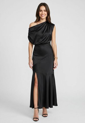 Femme souriante portant une robe en satin noire sans manches, avec une épaule dénudée et une fente devant, assortie à des talons noirs à bride à la cheville.