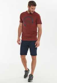 Bordeauxrotes T-Shirt mit navyblauem grafischem Design, kombiniert mit navyblauen Shorts. Model trägt graue Sneakers und zeigt einen lässigen, sportlichen Look.