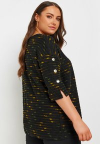 Svart texturerad topp med gula och gröna ränder, trekvartsärmar med knappdetaljer, rundad halsringning och avslappnad passform.