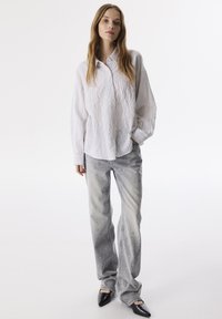 Camisa blanca texturizada de botones combinada con jeans grises que presentan un sutil degradado y adornados con tachuelas metálicas, completada con tacones negros.