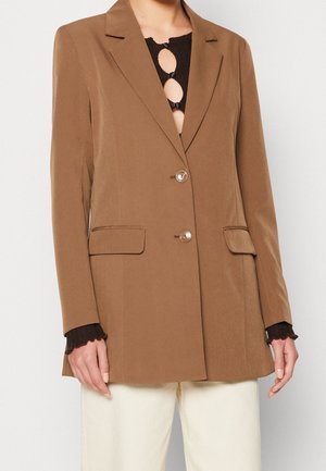 Lichtbruine dubbelgeknoopte blazer gedragen over een zwarte top met ronde uitsnijdingen en witte broek, getoond van nek tot heupen.