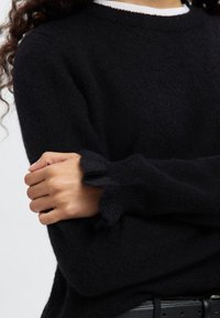 Svart stickad tröja med rund halsringning och volangkantade ärmslut, som visar en mjuk textur. Designen har en figurnära silhuett.