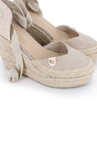 Cuñas de esparto beige con parte superior de tela texturizada, suela de yute entrelazada y lazos de cinta a juego. Presenta un detalle del logo en dorado.
