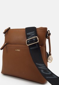 Borsa a tracolla in pelle marrone con texture, dotata di cinturino nero regolabile, tasca frontale con zip, logo dorato e dettaglio a forma di charm dorato circolare.