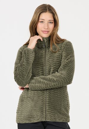 Junge Frau mit langen Haaren, die eine olivgrüne, strukturierte Fleecejacke mit Reißverschluss trägt und den Reißverschluss am Kragen hält, vor einem schlichten Hintergrund stehend.