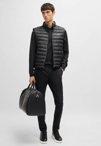 Black quilted vest over a black long-sleeve polo, paired with black pants and black sneakers. Model holds a black leather duffel bag.