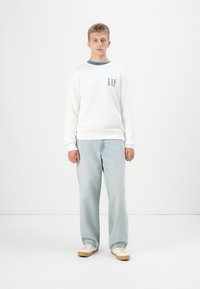 Sudadera blanca con acentos azules, que presenta un logo en el bolsillo. Combinada con jeans de mezclilla azul claro. El modelo lleva zapatillas blancas con suelas beige.