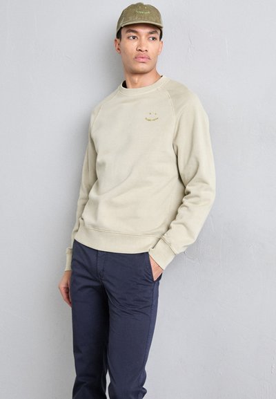 Lys beige genser med rund hals,mansjettærmer og en subtil logo. Mørkeblå bukser. Glatt tekstur, avslappet passform.