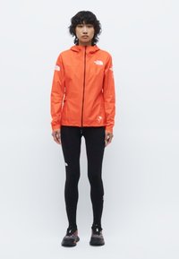Veste légère orange vif avec capuche, fermeture éclair à l'avant et logos sur les manches. Portée sur des leggings noirs et des chaussures de sport.