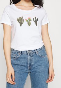 White short-sleeve t-shirt featuring printed cacti designs in green and pink. Light fabric with a round neckline and relaxed fit.