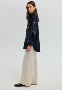 Camicia blu navy con ricami floreali, maniche lunghe e dettagli con bottoni, abbinata a pantaloni larghi crema e ballerine nere.