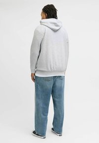 Grijze hoodie met een relaxed fit en een grote capuchon, gecombineerd met lichtblauwe baggy jeans. Het model draagt zwart-witte sneakers.