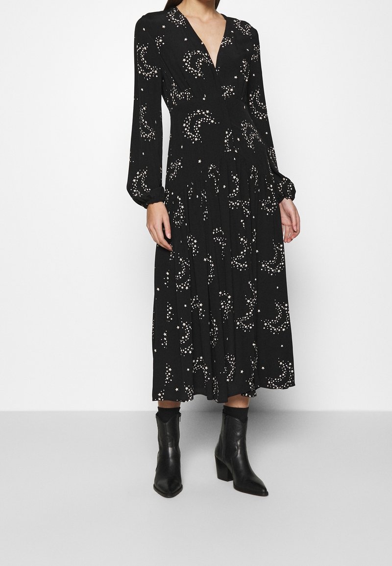 Femme portant une robe midi noire à manches longues avec des motifs d'étoiles et de lunes blanches et des bottines noires sur un fond uni.