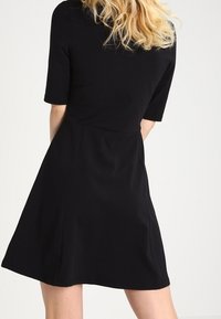 Svart klänning med korta ärmar och en figursydd kropp, som vidgar sig till en knä-lång fåll. Slät textur utan synliga mönster eller detaljer.