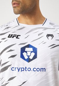 Camiseta atlética blanca con un diseño texturizado y detalles en negro, un logo hexagonal azul y el texto "crypto.com" destacado de manera prominente.