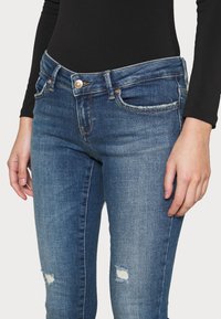 Jeans de mezclilla azul ajustados con un diseño desgastado, con desgarros visibles, cierre de botón y bolsillos en la parte delantera y trasera.