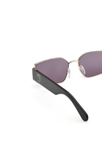 Gafas de sol angulares con lentes tintados en púrpura, acentos de marco de metal dorado, y patillas de acetato negro que presentan un detalle de logo.