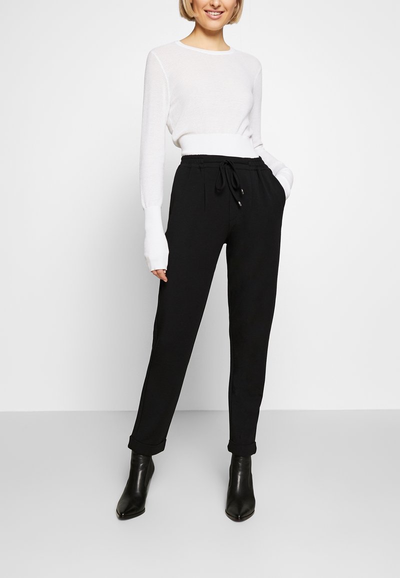 Pantalon de jogging noir avec taille à cordon, poches latérales et jambes fuselées, associé à un haut blanc ajusté à manches longues.