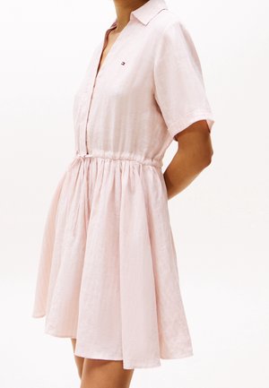 Femme portant une robe rose clair, à manches courtes, avec un col et une taille froncée, debout sur un fond blanc uni.