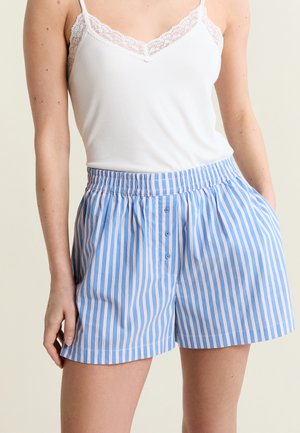 Frau trägt ein weißes Tanktop mit Spitzenbesatz und blau-rosa vertikal gestreifte Shorts mit Knöpfen und elastischem Bund, steht mit der Hand in der Tasche.