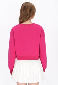 Rózsaszín, rövidített fazonú pulóver hosszú ujjakkal, kerek nyakkivágással és bordázott szegéllyel. Alatta fehér, rakott szoknyával párosítva.