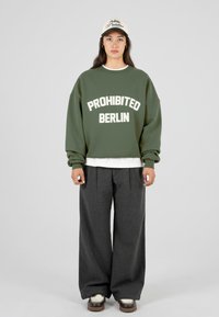 Jauna moteris stovi vilkėdama alyvuogių spalvos džemperį su užrašu „PROHIBITED BERLIN“, plačias tamsias kelnes, baltus sportinius batelius ir smėlio spalvos kepurę su juodu tekstu.