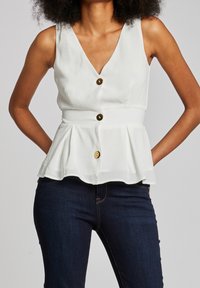 Bílý peplum top s véčkovým výstřihem, třemi velkými hnědými knoflíky vpředu a strukturovanou tkaninou. Skombinováno se tmavě modrými džíny.