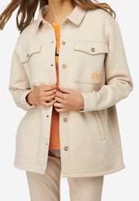 Beige-colourinen nappikiinnitteinen takki, jossa on kaksi rintataskua, pyöristetty helma ja oranssi kirjailu. Päällä raidallinen oranssin ja sinisen yhdistelmäpaita.