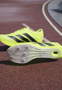 Bright yellow athletic shoes with black three-stripe design, textured upper, and a white outsole with six metal spikes and a visible logo.