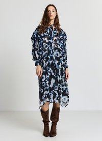 Fließendes, langes dunkelblaues Kleid mit einem Blumenmuster in verschiedenen Blautönen, versehen mit gerafften Ärmeln und einem Bindeband am Ausschnitt, kombiniert mit braunen Stiefeln.